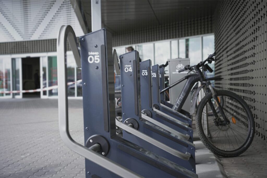 Racks de stationnement intelligents Bikeep numérotés, avec un VTT électrique noir garé à proximité de l'entrée d'un bâtiment.