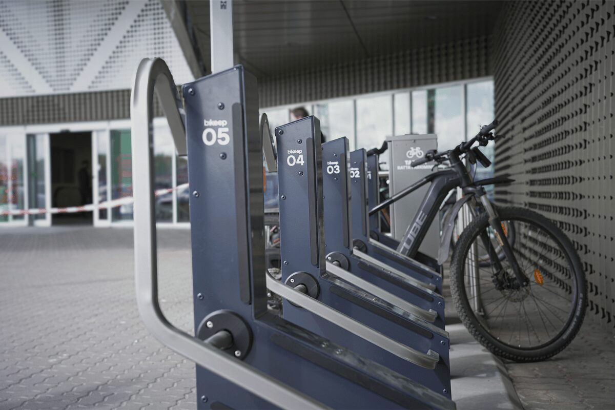 Racks de stationnement intelligents Bikeep numérotés, avec un VTT électrique noir garé à proximité de l'entrée d'un bâtiment.